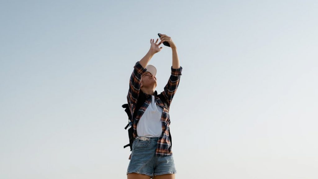 A young woman in casual attire taking a selfie against a clear sky.